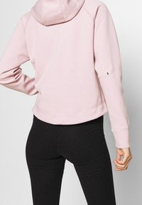 Felpa rosa con cappuccio dal taglio rilassato, caratterizzata da maniche raglan e orlo a costine, abbinata a leggings neri a righe verticali.