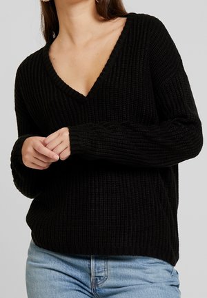 Femme portant un pull noir en maille côtelée à col en V et un jean bleu clair, les mains délicatement jointes devant elle, fond neutre.