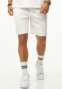 Weiße strukturierte Shorts mit einem Rautenmuster, umgeschlagenem Saum, kombiniert mit weißen Sneakers und weißen Socken mit schwarzen Streifen.