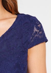 Námořnická modrá krajková blůza s krátkými rukávy, která má texturovaný květinový vzor a jemný, průhledný výstřih.