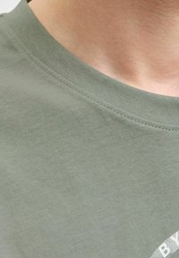 Camiseta de algodón verde oliva con cuello redondo, que presenta una textura suave y un detalle de logo impreso en blanco en la parte inferior derecha.