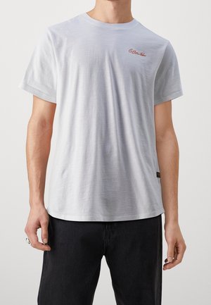 Mężczyzna ubrany w gładki biały T-shirt z krótkim rękawem z małym czerwonym napisem na lewej piersi i czarne spodnie na jednolitym tle.