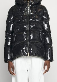 Černá lesklá péřová bunda s předním zipem a štítkem "PINKO" na poutku zipu, nošená s bílými kalhotami viditelnými od pasu dolů.