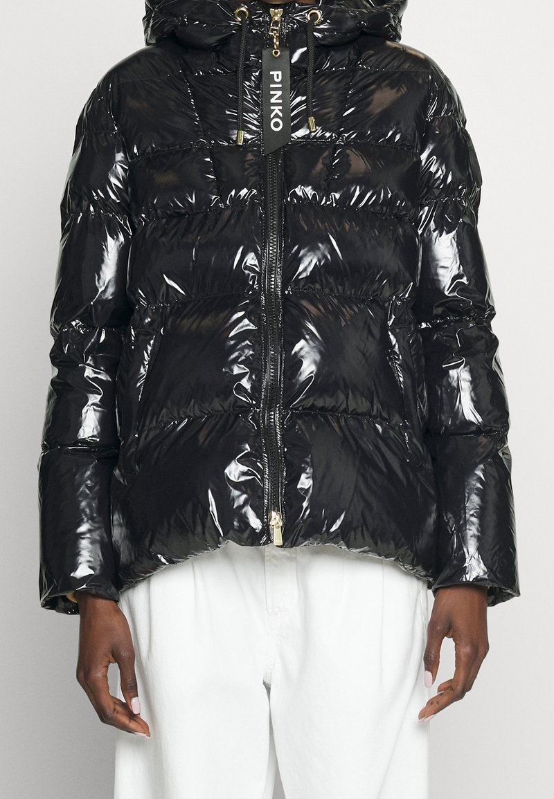 Černá lesklá péřová bunda s předním zipem a štítkem "PINKO" na poutku zipu, nošená s bílými kalhotami viditelnými od pasu dolů.