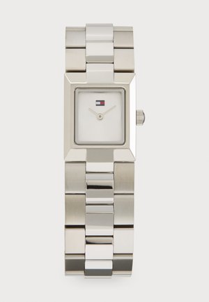 Ezüst színű rozsdamentes acél óra négyzet alakú számlappal, fehér tárcsával, vékony mutatókkal és egy kis logóval az óránként 12-nél. Lánckarkötő kivitel.