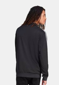 Chaqueta deportiva negra con un alto cuello, tejido texturizado y rayas blancas a lo largo de los hombros. Presenta un corte ajustado y bolsillos con cremallera.