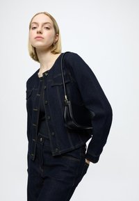 Σκούρο τζιν μπουφάν με χρυστά ραφές και τσέπες, συνδυασμένο με ταιριαστό παντελόνι. Μικρή μαύρη τσάντα φορεμένη στον ώμο.