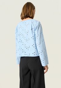 Ljusblå, kort blus med lång ärm, som har öppet arbete detaljer och en texturerad yta. Designad med en rund halsringning.