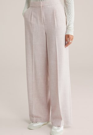 Kvinde iført højtaljede, vidde, beige hørbukser med sidelommer, kombineret med hvide sneakers og en cremet tekstureret langærmet top.