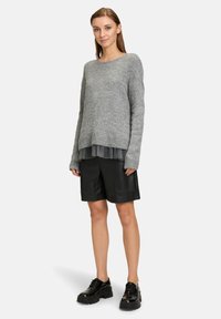 Pull en maille gris avec un large col, superposé sur un haut court, transparent et plissé ; associé à un short en cuir noir et des chaussures noires épaisses.