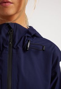 Chaqueta azul marino resistente al agua con un alto cuello, que presenta un cierre frontal con cremallera y toggles ajustables. Textura suave con detalles de hardware mínimos.