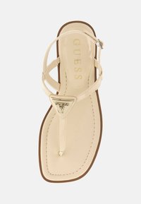 Beige tanga sandal med en glänsande yta, en triangelformad metallisk logotypaccent och en platt brun sula med sömdetalj.