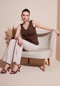 Brown sleeveless top, light beige flared pants, and brown braided high-heeled sandals. Sitting on a light grey, upholstered chair with buttons.