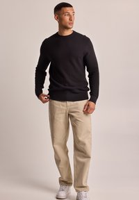 Black textured knit sweater with a round neckline, paired with light beige pants and white sneakers. The fit is casual and relaxed.