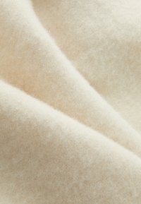 Tissu en laine beige doux avec une texture fine et duveteuse, présentant de doux plis qui créent des ombres subtiles et de la profondeur dans le matériau.