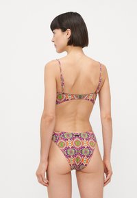 Vrouw met kort donker haar, die een bikini met paars, oranje en groen patroon draagt, staande met de rug naar de camera tegen een witte achtergrond.