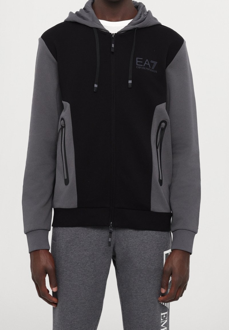 Man som bär en svart och grå zip-up hoodie med huva och snören samt gråa sweatpants med sidotryck.