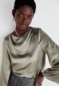 Olive green, draped blouse with a soft, shiny texture and wide sleeves, paired with a gray plaid skirt. Silver chain necklace and earrings.