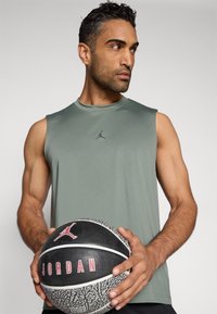 Πράσινο αμάνικο αθλητικό μπλουζάκι με διακριτικό λογότυπο, κρατούμενο από άτομο που κρατάει μια μαύρη και γκρι μπάλα μπάσκετ με κόκκινες λεπτομέρειες και λογότυπο.