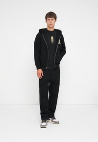 Black zip-up hoodie over a black t-shirt with graphic, paired with black cargo pants and white sneakers with gray and yellow accents.