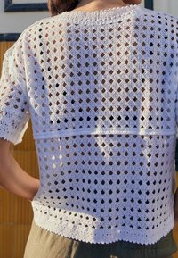 Bílý háčkovaný top se vzorem otevřeného diamantu, krátkými rukávy, žebrovaným výstřihem a rovním spodním lemem, odhalující kůži pod ním.