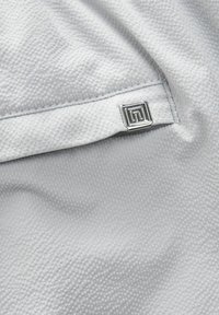 Grau textil mit einer subtil gemusterten Oberfläche und einem silbernen, geometrischen Logo-Tag, das in der unteren rechten Ecke angebracht ist.