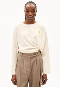 Beiges langärmeliges Baumwollhemd mit lockerem Schnitt und gelbem Banddetail; kombiniert mit hochtaillierter, beige plissierter Hose.