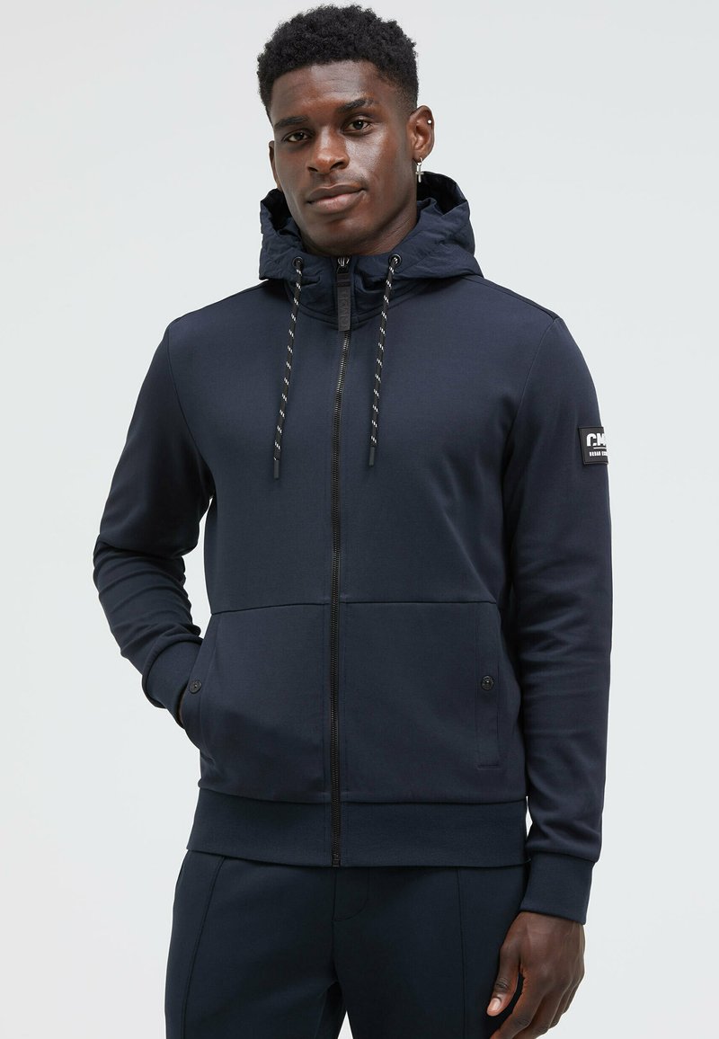 Navy zip-up hoodie made of smooth fabric, featuring a drawstring hood, side pockets, and a logo patch on the left arm.