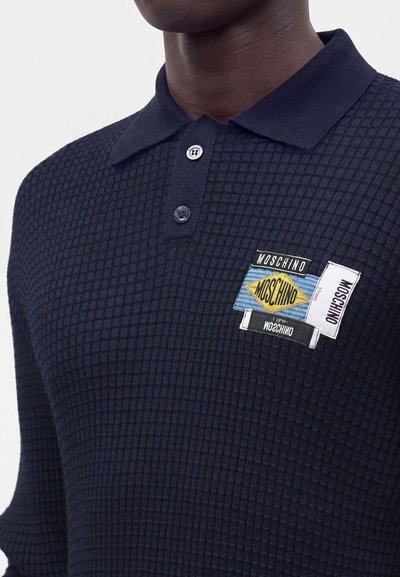 Polo à manches longues texturé bleu foncé avec deux boutons et plusieurs patches logo Moschino sur la poitrine gauche.