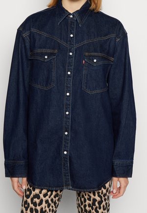 Mørkeblå denimskjorte med knapper og klaplommer båret over beige og sorte bukser med leopardprint.