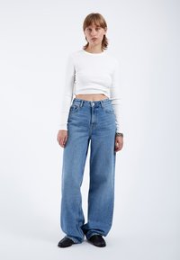 White long-sleeve cropped top paired with high-waisted, loose-fitting light blue denim jeans and black textured footwear.