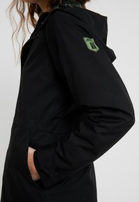 Giacca nera impermeabile con cappuccio, caratterizzata da un logo verde sulla manica e una superficie liscia e strutturata. Il design prevede un punto vita arricciato.