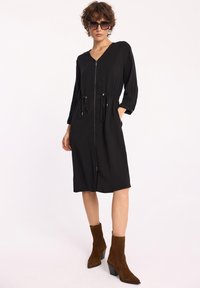 Czarna sukienka z dekoltem w kształcie litery V, zamek z przodu i ściągacz w pasie. Posiada rękawy o długości 3/4 oraz boczne kieszenie. Zestawiona z brązowymi botkami za kostkę.