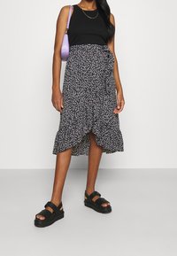 Top senza maniche nero abbinato a una gonna avvolgente floreale, lunga fino al ginocchio, con orlo arricciato e accenti rosa. Sandali con piattaforma neri completano il look.