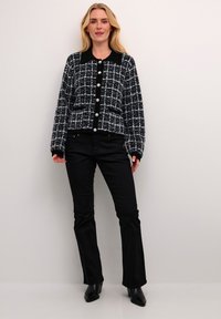 Melnā un baltā šaha modeļa kardigāns ar pērļu pogām, melnu apkakli un ribotu apakšmalu, kombinēts ar melniem platuma biksēm un norādītām zābakām.