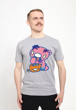 Mand med overskæg iført en grå t-shirt med en farverig tegneseriebamse, der slikker på et dryppende orange objekt, stående mod en hvid baggrund.