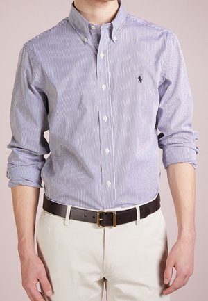 Hombre con camisa de botones de manga larga a rayas azules y blancas, con las mangas remangadas, pantalones beige y cinturón negro.