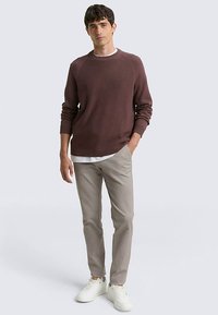 Pull côtelé marron avec col rond, associé à un pantalon beige clair et des baskets blanches. Le modèle pose les mains dans les poches devant un fond neutre.