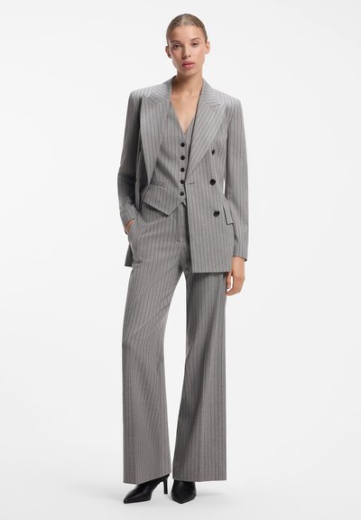 Femme portant un costume trois pièces gris à fines rayures avec un blazer croisé, un gilet, un pantalon à jambes larges et des escarpins noirs à bouts pointus.
