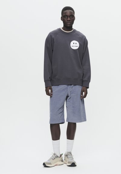 Grauer Pullover mit einem runden, weißen Gesichtsgrafik, kombiniert mit blauen Cordshorts. Das Model trägt weiße Socken und beigen Sneakers.