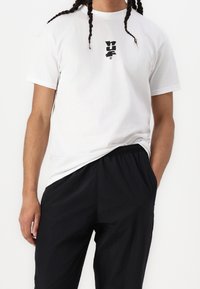 Bílý bavlněný tričko s černým logem na hrudi, v kombinaci s černými kalhotami s elastickým pasem. Tkanina se zdá být hladká a lehká.