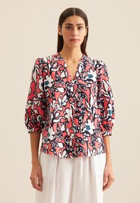 Kobieta w czerwono-czarno-biało-niebieskiej bluzce w kwiatowy wzór z rękawami do łokci i białych spodniach, stojąca na tle neutralnym.