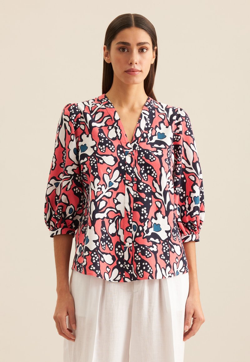 Kobieta w czerwono-czarno-biało-niebieskiej bluzce w kwiatowy wzór z rękawami do łokci i białych spodniach, stojąca na tle neutralnym.