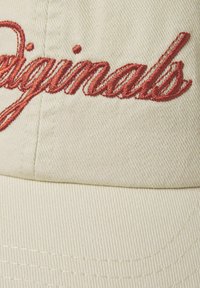 Beige bomulds cap med rød broderet "originals" tekst. Teksten har et flydende skriftdesign med synlige sømdetaljer.