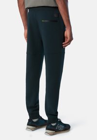 Pantalones jogger verde oscuro con corte ajustado, que cuentan con un bolsillo trasero con cremallera y detalles de costura en contraste, combinados con zapatillas deportivas negras.