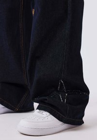 Baskets blanches partiellement recouvertes par un jean bleu foncé ample avec des coutures orange, posées sur une surface claire.
