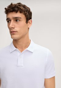 Witte polo met een klassieke kraag en twee knopen. Kenmerkt zich door een textuurstof en een ingekleurde logo op de borst. Eenvoudig en strakke ontwerp.