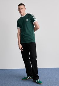 Žalios medvilnės marškinėliai su baltais akcentais ir Adidas logotipu, su juodomis kelnėmis ir žaliais batais su baltais dryžiais. Modelis stovi su viena ranka už nugaros.
