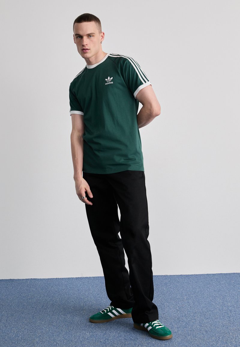 T-shirt en coton vert avec des bordures blanches et le logo Adidas, associé à un pantalon noir et des chaussures vertes avec des rayures blanches. Le modèle se tient avec une main derrière le dos.