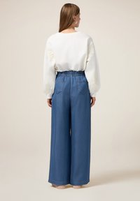 Sweatshirt blanc avec des manches texturées et une taille élastique, associé à un pantalon en denim bleu à jambes larges comprenant deux poches arrière et une coupe décontractée.